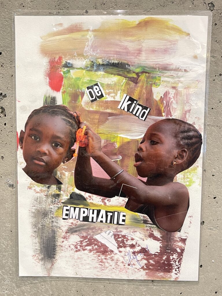 Die Collage von Daniela Hafner zeigt zwei dunkelhäutige Mädchen. Eines steckt dem anderen Blumen ins Haar. Mit Zeitungslettern aufgeklebt ist der Text "Be kind - Empathie" zu lesen.